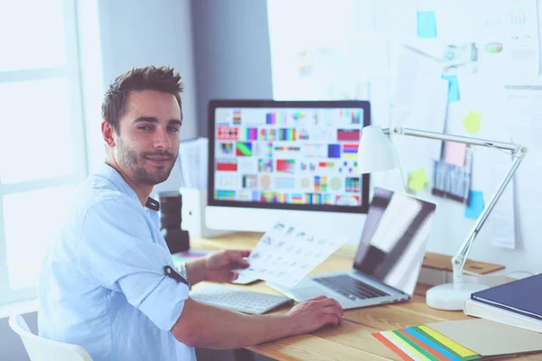 Retrato del joven diseñador sentado en el estudio gráfico frente a la computadora portátil y el ordenador mientras trabaja en línea. — Foto de Stock