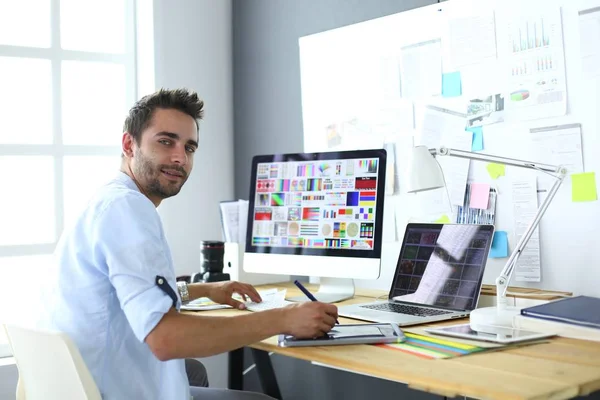 Retrato del joven diseñador sentado en el estudio gráfico frente a la computadora portátil y el ordenador mientras trabaja en línea. — Foto de Stock