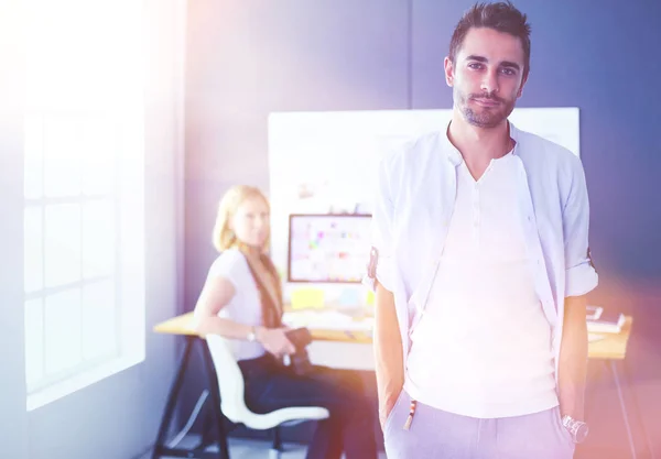 Porträt eines jungen Designers vor Laptop und Computer während der Arbeit. Assistentin bedient ihr Handy im Hintergrund. — Stockfoto