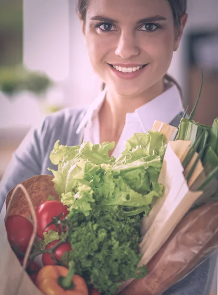 Mujer joven sosteniendo bolsa de la compra de comestibles con verduras de pie en la cocina. —  Fotos de Stock