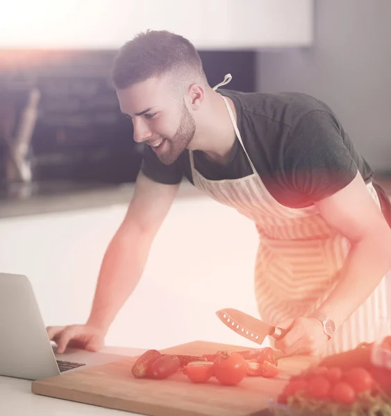 Porträt eines Mannes, der Gemüse in der Küche kocht, während er auf einen Laptop auf dem Tisch blickt — Stockfoto