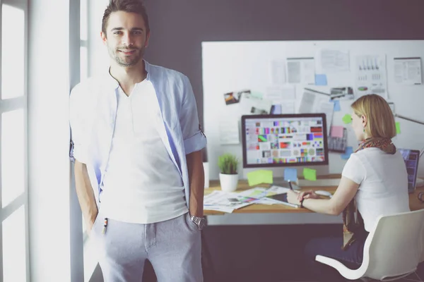 Porträt eines jungen Designers vor Laptop und Computer während der Arbeit. Assistentin bedient ihr Handy im Hintergrund. — Stockfoto