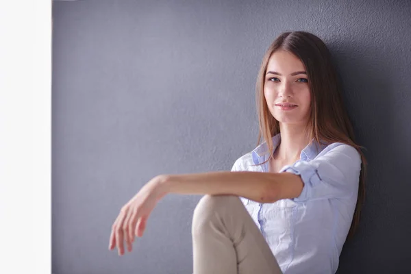 Retrato de una mujer feliz y casual sentada en el suelo sobre fondo gris —  Fotos de Stock