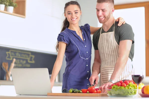 Jovem cortando legumes e mulher de pé com laptop na cozinha — Fotografia de Stock