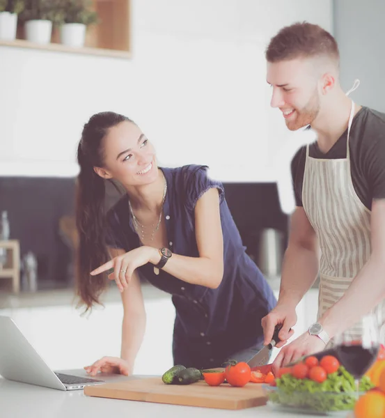 Hombre joven cortando verduras y mujer de pie con el ordenador portátil en la cocina —  Fotos de Stock