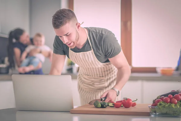 Hombre joven cortando verduras y mujer de pie con el ordenador portátil en la cocina — Foto de Stock