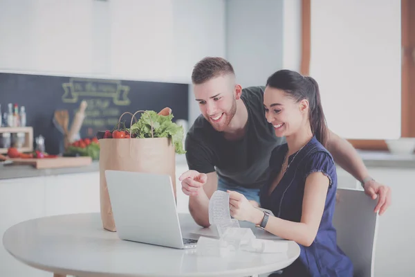 Jongeman snijden groenten en vrouw staan met laptop in de keuken — Stockfoto