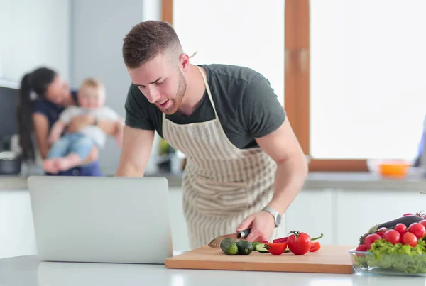 Jongeman snijden groenten en vrouw staan met laptop in de keuken — Stockfoto