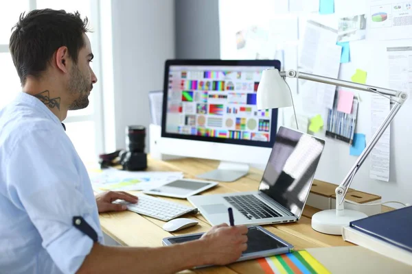 Retrato del joven diseñador sentado en el estudio gráfico frente a la computadora portátil y el ordenador mientras trabaja en línea. — Foto de Stock