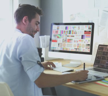 İnternette çalışırken bilgisayarın ve dizüstü bilgisayarın önünde grafik stüdyosunda oturan genç tasarımcının portresi.