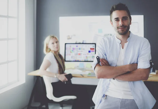 Porträt eines jungen Designers vor Laptop und Computer während der Arbeit. Assistentin bedient ihr Handy im Hintergrund. — Stockfoto