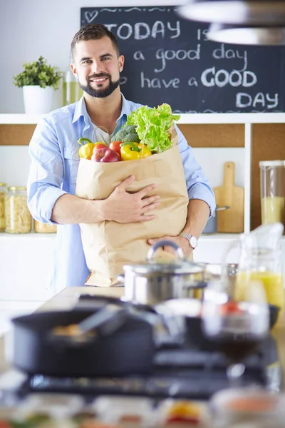 Facet z papierową torbą pełną zakupów na tle kuchni. Zakupy i koncepcja zdrowej żywności — Zdjęcie stockowe