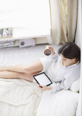 Sabahları yatakta otururken tablet kullanan ve kahve içen kadın portresi