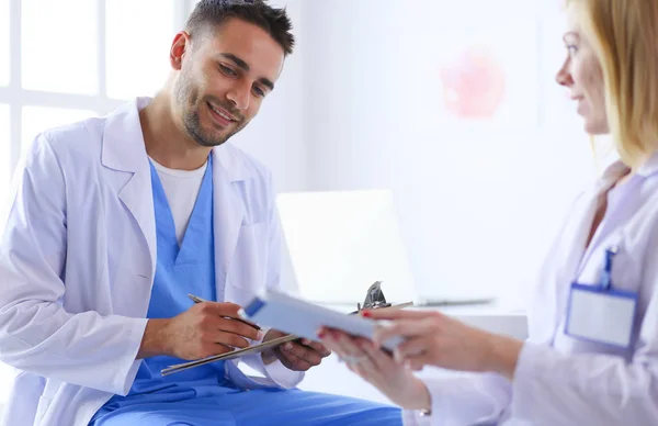 Médico bonito está conversando com jovem médica e fazendo anotações enquanto está sentado em seu escritório. — Fotografia de Stock