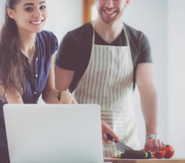 Sebze doğrayan genç bir adam ve mutfakta dizüstü bilgisayarı olan bir kadın.