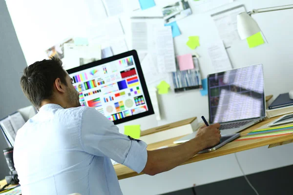 Retrato del joven diseñador sentado en el estudio gráfico frente a la computadora portátil y el ordenador mientras trabaja en línea. — Foto de Stock