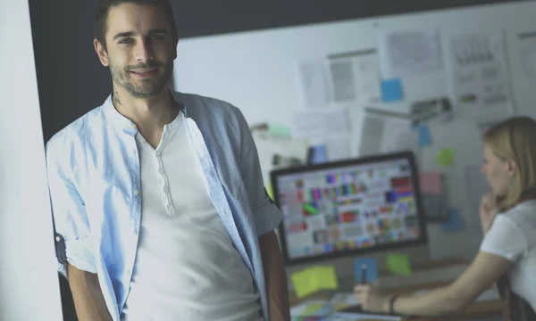 Retrato del joven diseñador delante de la computadora portátil y el ordenador mientras trabaja. Asistente usando su móvil en segundo plano . — Foto de Stock