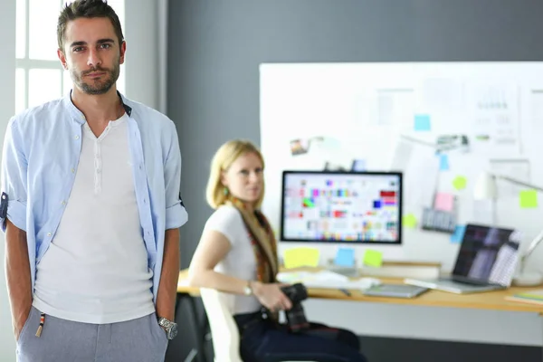 Porträt eines jungen Designers vor Laptop und Computer während der Arbeit. Assistentin bedient ihr Handy im Hintergrund. — Stockfoto