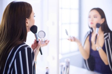 Güzel kız aynaya bakıyor ve büyük bir fırçayla kozmetik uyguluyor.