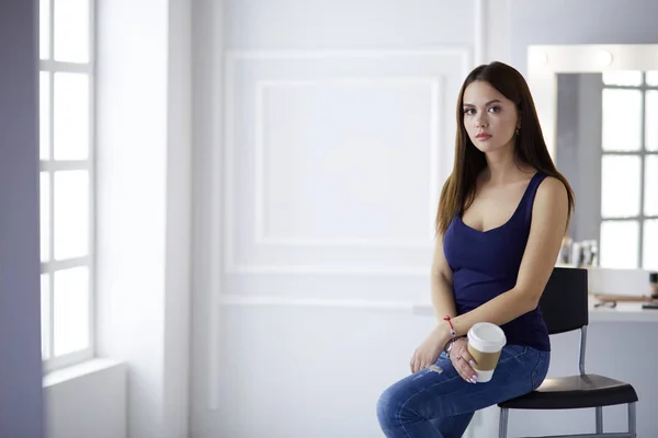 Bella giovane donna che tiene in mano la tazza di caffè e distoglie lo sguardo con il sorriso mentre si siede sulla sedia — Foto Stock