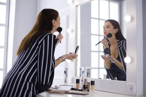Attractive young woman doing make-up while looking at the mirror and smiling