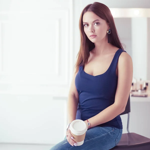 Hermosa mujer joven sosteniendo la taza de café y mirando hacia otro lado con sonrisa mientras está sentado en la silla —  Fotos de Stock