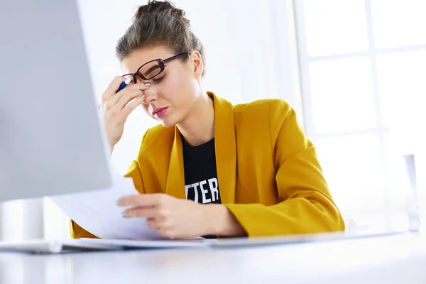 Attractive woman sitting at desk in office, working with laptop computer, holding document — Stock Photo, Image