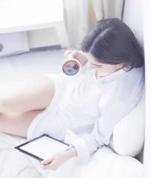 Portrait of woman using tablet and drinking coffee while sitting on the bed in the morning — Stock Photo, Image