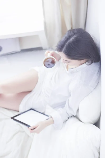 Portret van een vrouw die een tablet gebruikt en 's ochtends koffie drinkt terwijl ze op bed zit — Stockfoto