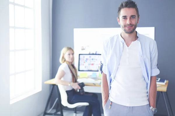 Porträt eines jungen Designers vor Laptop und Computer während der Arbeit. Assistentin bedient ihr Handy im Hintergrund. — Stockfoto