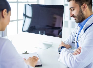 Yakışıklı doktor genç bayan hastasıyla konuşuyor ve ofisinde otururken notlar alıyor.