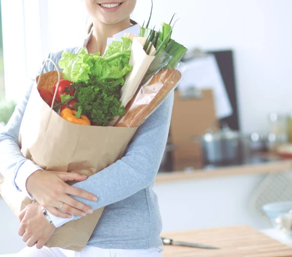 Mujer joven sosteniendo bolsa de la compra de comestibles con verduras. De pie en la cocina —  Fotos de Stock