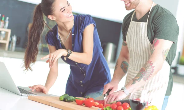 Hombre joven cortando verduras y mujer de pie con el ordenador portátil en la cocina — Foto de Stock