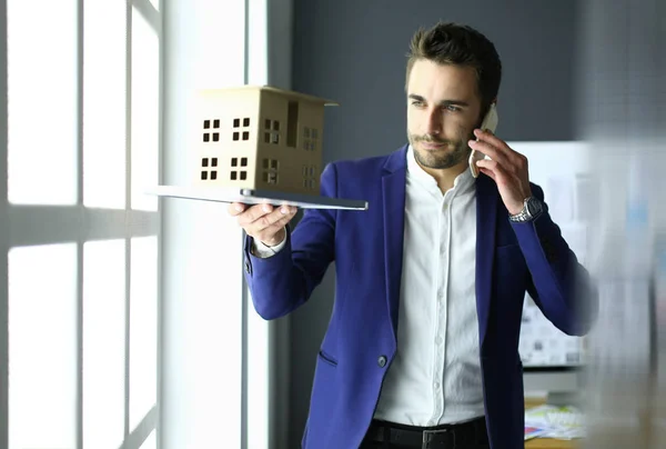 Empresário segurando casa em miniatura na mão de pé no escritório . — Fotografia de Stock