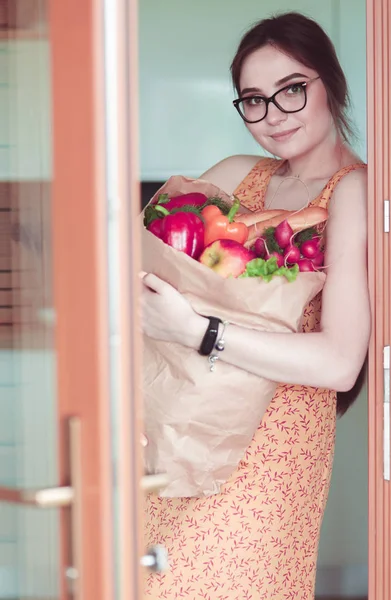 Mujer joven sosteniendo bolsa de la compra de comestibles con verduras. De pie en la cocina. —  Fotos de Stock