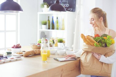 Sebzeli alışveriş poşeti taşıyan genç bir kadın. Mutfakta duruyor.