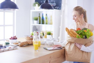 Sebzeli alışveriş poşeti taşıyan genç bir kadın. Mutfakta duruyor.