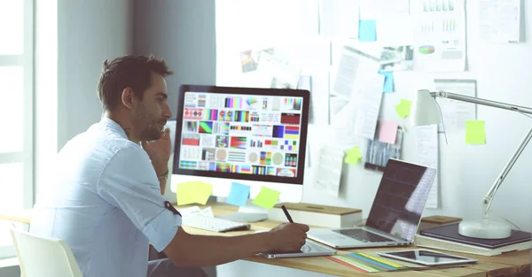 Retrato del joven diseñador sentado en el estudio gráfico frente a la computadora portátil y el ordenador mientras trabaja en línea. — Foto de Stock