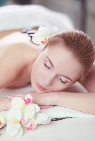 Mujer joven disfrutando de un masaje de roca caliente en un salón de spa y flor blanca en su cabeza — Foto de Stock