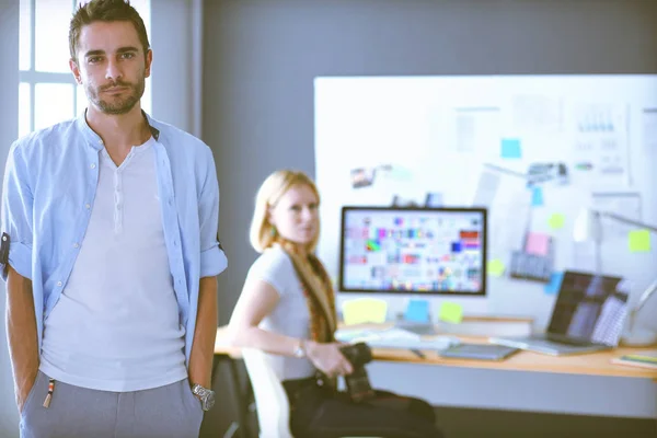 Porträt eines jungen Designers vor Laptop und Computer während der Arbeit. Assistentin bedient ihr Handy im Hintergrund. — Stockfoto