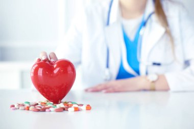 Steteskoplu bir doktor, beyaz arka planda kırmızı kalbi inceliyor.