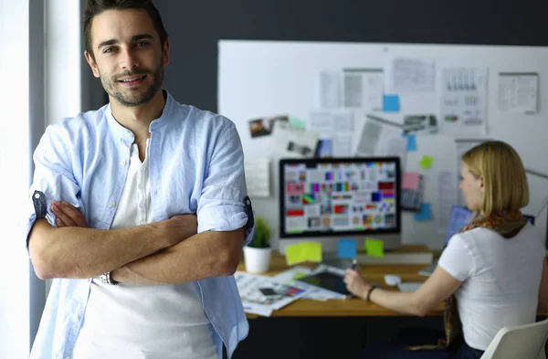 Porträt eines jungen Designers vor Laptop und Computer während der Arbeit. Assistentin bedient ihr Handy im Hintergrund. — Stockfoto