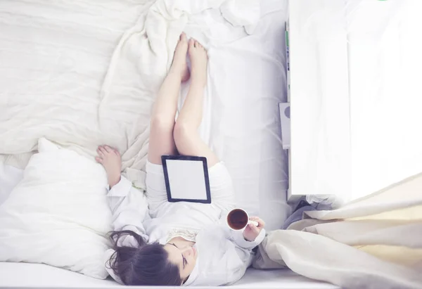 Portrait of woman using tablet and drinking coffee while sitting on the bed in the morning — Stock Photo, Image