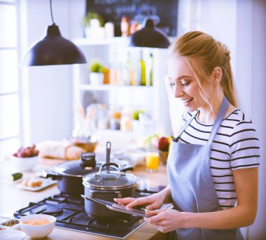 Mutfakta yemek yapmak için tablet bilgisayar kullanan genç bir kadın.