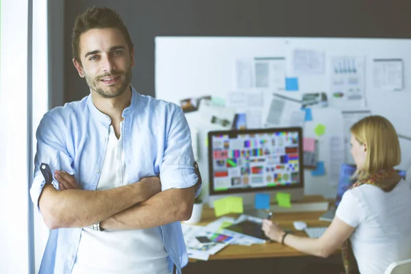 Porträt eines jungen Designers vor Laptop und Computer während der Arbeit. Assistentin bedient ihr Handy im Hintergrund. — Stockfoto