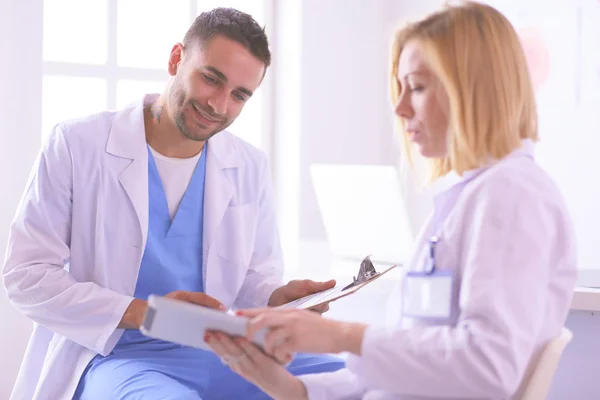 Médico bonito está conversando com jovem médica e fazendo anotações enquanto está sentado em seu escritório. — Fotografia de Stock