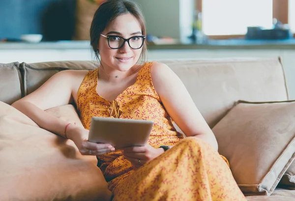 Hermosa joven morena en casa sentada en un sofá o sofá usando su tablet y sonriendo — Foto de Stock