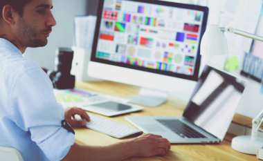 İnternette çalışırken bilgisayarın ve dizüstü bilgisayarın önünde grafik stüdyosunda oturan genç tasarımcının portresi.