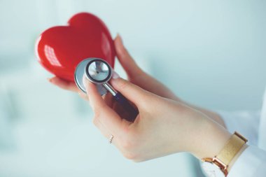 Steteskoplu bir doktor, beyaz arka planda kırmızı kalbi inceliyor.