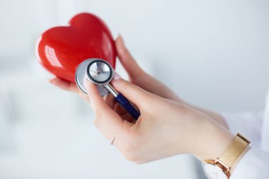 Steteskoplu bir doktor, beyaz arka planda kırmızı kalbi inceliyor.